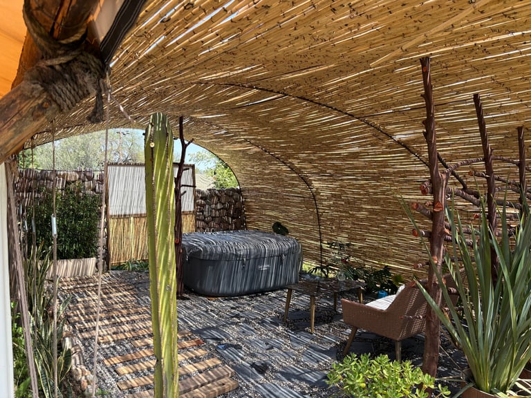 Rustic outdoor patio with woven reed ceiling, vintage metal bathtub, wooden furniture, potted plants, and warm lighting at sunset