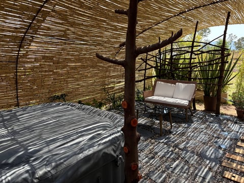 Rustic outdoor patio with thatched roof, wooden bed frame on left, and white cushioned bench with decorative branches on right